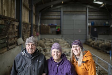 Three people, two elderly and one younger, wearing winter clothing and knit caps, pose inside a barn with sheep in the background.