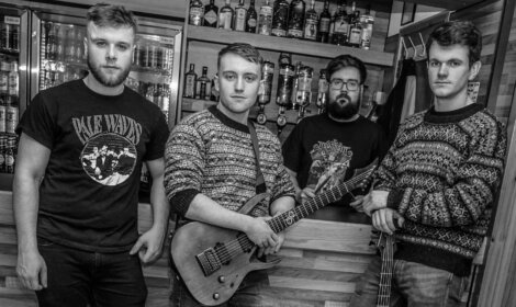 A black and white photo of four men in a room with shelves of bottles. The two in front hold guitars, one wears a band t-shirt, and the others wear patterned sweaters.