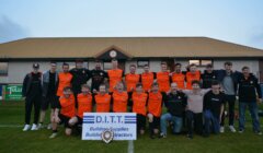A group of soccer players in orange jerseys and others in casual attire pose together outdoors in front of a building, with a sign that reads "D.I.T.T. Building Supplies Building Contractors.