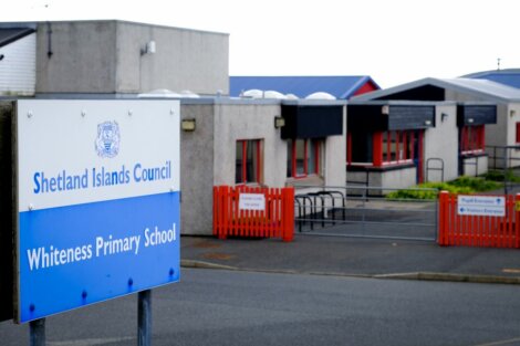 Sign reading "Shetland Islands Council Whiteness Primary School" in front of a school building with red fencing and blue trim.