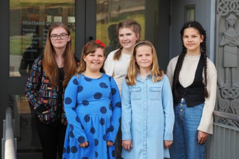 Five girls standing in front of a building entrance, smiling at the camera. The building door has visible text indicating hours of operation. Two statues are carved into the wall on the right.