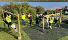A group of people, some in high-visibility clothing, gather around a wooden outdoor exercise structure by a river. Some are using the equipment, while others stand nearby.