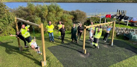 A group of people, some in high-visibility clothing, gather around a wooden outdoor exercise structure by a river. Some are using the equipment, while others stand nearby.