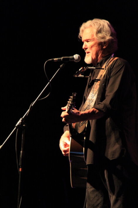 A man with gray hair is standing on a dark stage, playing an acoustic guitar and singing into a microphone. He has a harmonica holder around his neck.
