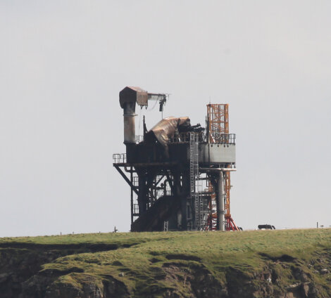 An industrial structure with metallic components stands on a grassy landscape, set against a cloudy sky.