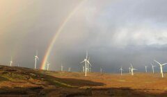 Wind turbines on a grassy landscape with a rainbow in the cloudy sky.