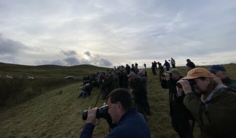 A large group of people are gathered on a grassy hillside, observing with binoculars and cameras under a cloudy sky.