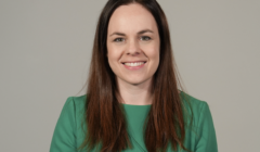 A person with long brown hair, wearing a green top, smiles with a neutral background.