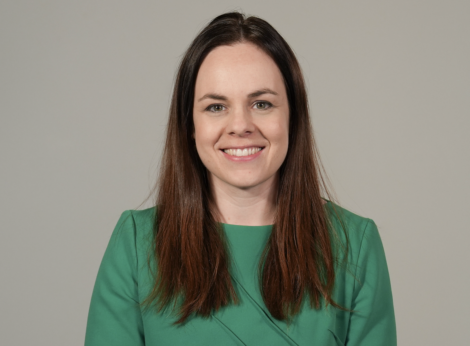 A person with long brown hair, wearing a green top, smiles with a neutral background.