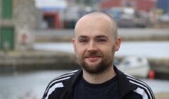 A man with a beard and a shaved head smiles slightly, standing outdoors near water with boats and buildings in the background.