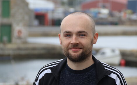 A man with a beard and a shaved head smiles slightly, standing outdoors near water with boats and buildings in the background.