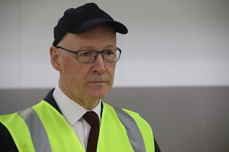 A man wearing glasses, a black cap, and a neon yellow safety vest with reflective strips stands in a neutral setting.