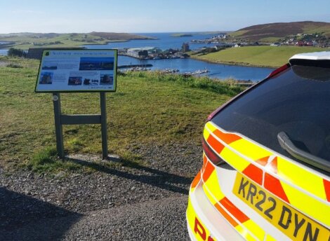 A police car is parked on a scenic overlook with a view of a coastal town and body of water. An informational sign is mounted nearby.
