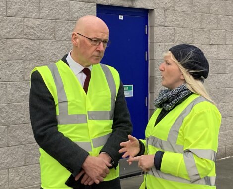 Two people in yellow safety vests and hairnets stand in front of a gray brick wall and blue door, engaged in conversation.