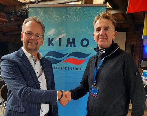 Two men shaking hands in front of a "KIMO Nederland en België" banner, one wearing a striped suit and the other in a black jacket.