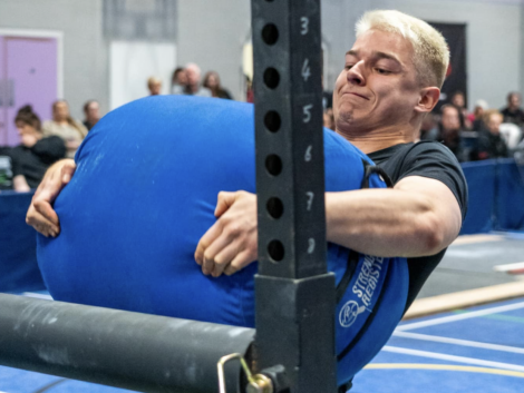 Person lifting a large blue sandbag during a competition, with spectators in the background.