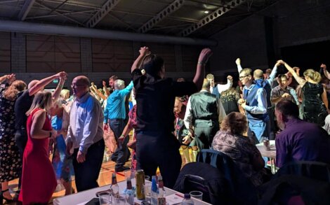 A group of people dancing energetically indoors, with others seated at tables front right. The room is dimly lit with a spacious ceiling.