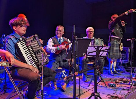 Three musicians play accordions and a guitar onstage while a person in a kilt speaks into a microphone.