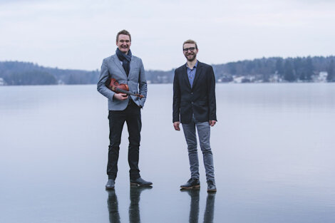 Two men stand on an icy surface; one holds a violin. Trees and water are visible in the background.