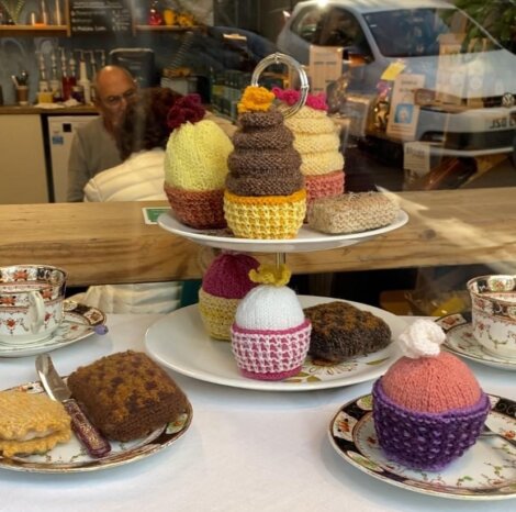 Display of knitted cupcakes on a tiered stand with teacups and saucers on the table, viewed through a window with a man and parked car in the background.