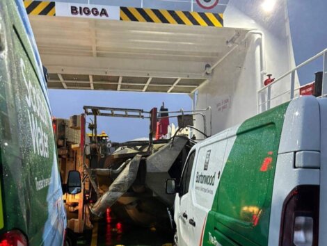 Vehicles tightly packed on a ferry deck, including two vans and a large piece of machinery, under wet and cloudy conditions.