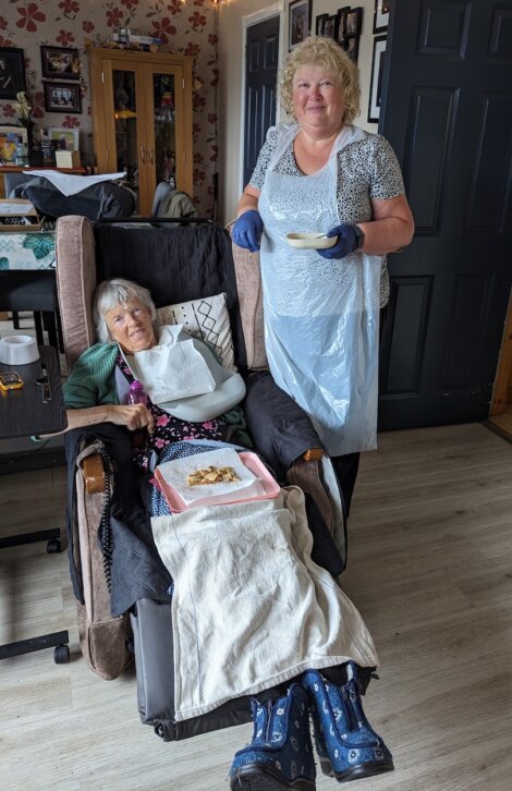 An elderly woman sits in a recliner with a meal tray, while another woman stands beside her holding a bowl and spoon, wearing gloves and an apron.