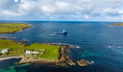 Aerial view of a blue ship navigating coastal waters near a grassy shoreline and rocky areas under a partly cloudy sky.