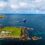 Aerial view of a blue ship navigating coastal waters near a grassy shoreline and rocky areas under a partly cloudy sky.
