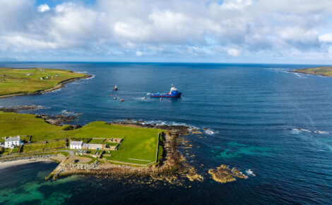 Aerial view of a blue ship navigating coastal waters near a grassy shoreline and rocky areas under a partly cloudy sky.