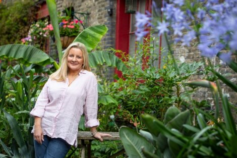 A person in a light pink shirt stands in a lush garden with colorful flowers and greenery, next to a brick building with a red door.