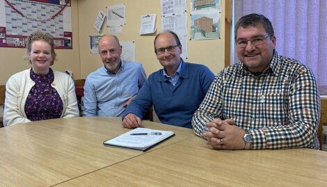 Four people sitting at a table with a notebook, smiling at the camera, in a room with maps and diagrams on the wall.