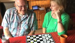 An elderly man and a woman smile while sitting at a table with a checkers board, next to two mugs, in a cozy room.