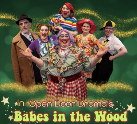 A group of six individuals in colorful costumes pose cheerfully in front of a green background with glittery accents. The text reads "Open Door Drama's Babes in the Wood.