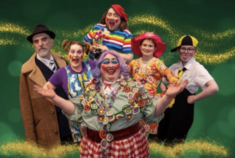 A group of six individuals in colorful costumes pose cheerfully in front of a green background with glittery accents. The text reads "Open Door Drama's Babes in the Wood.
