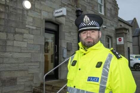 A police officer in a bright yellow uniform stands outside a stone police station building.