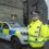 Police officer in a high-visibility jacket stands by two police vehicles outside a stone building.