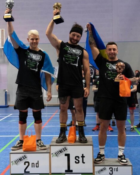 Three men stand on a podium. The first-place winner holds a trophy and is flanked by second and third place winners. They wear black shirts and hold flags.