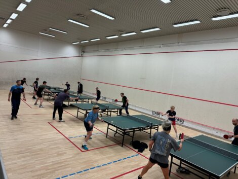 People playing table tennis in a large indoor room with multiple tables.