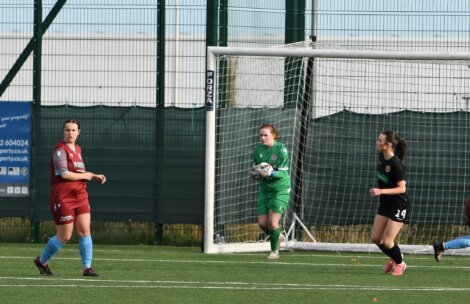 Soccer players on a field during a game, with a goalkeeper in green holding the ball near the goalpost. Two players in action, dressed in red and black uniforms.