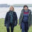 Two individuals walk on a grassy hill overlooking a beach and ocean under a cloudy sky.