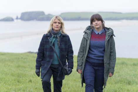 Two individuals walk on a grassy hill overlooking a beach and ocean under a cloudy sky.
