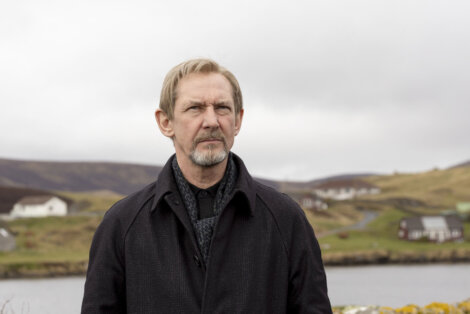 A man with short hair and a beard stands outdoors in front of a rural landscape with houses and hills under a cloudy sky.