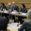 A group of people sit at a long table with laptops and microphones during a meeting in a conference room.