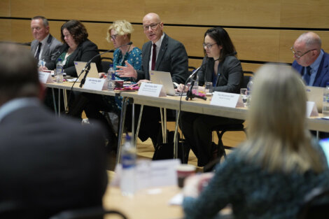 A group of people sit at a long table with laptops and microphones during a meeting in a conference room.