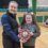 Two people smiling, one holding a shield-shaped award, standing in a gymnasium with empty seats in the background.