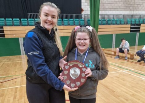 Two people smiling, one holding a shield-shaped award, standing in a gymnasium with empty seats in the background.