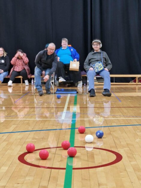 Two people play boccia indoors on a wooden floor, aiming for a white target ball. Spectators watch in the background.