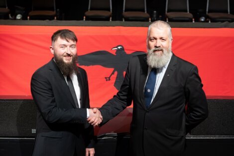 Two men in suits with beards shake hands in front of a red flag featuring a black raven.