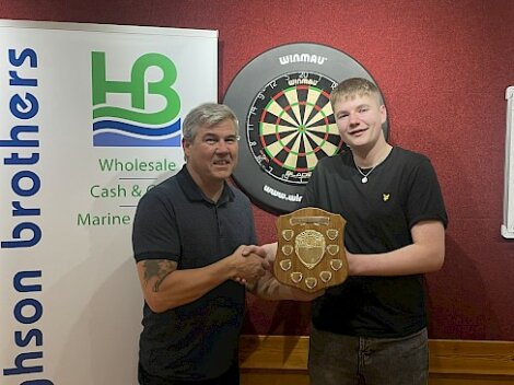 Two men shaking hands in front of a dartboard. One man holds a plaque. A sign on the left reads "Johnson Brothers Wholesale Cash & Marine.
