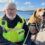 A man wearing a high-visibility vest sits on rocks by the sea with a dog on a leash.